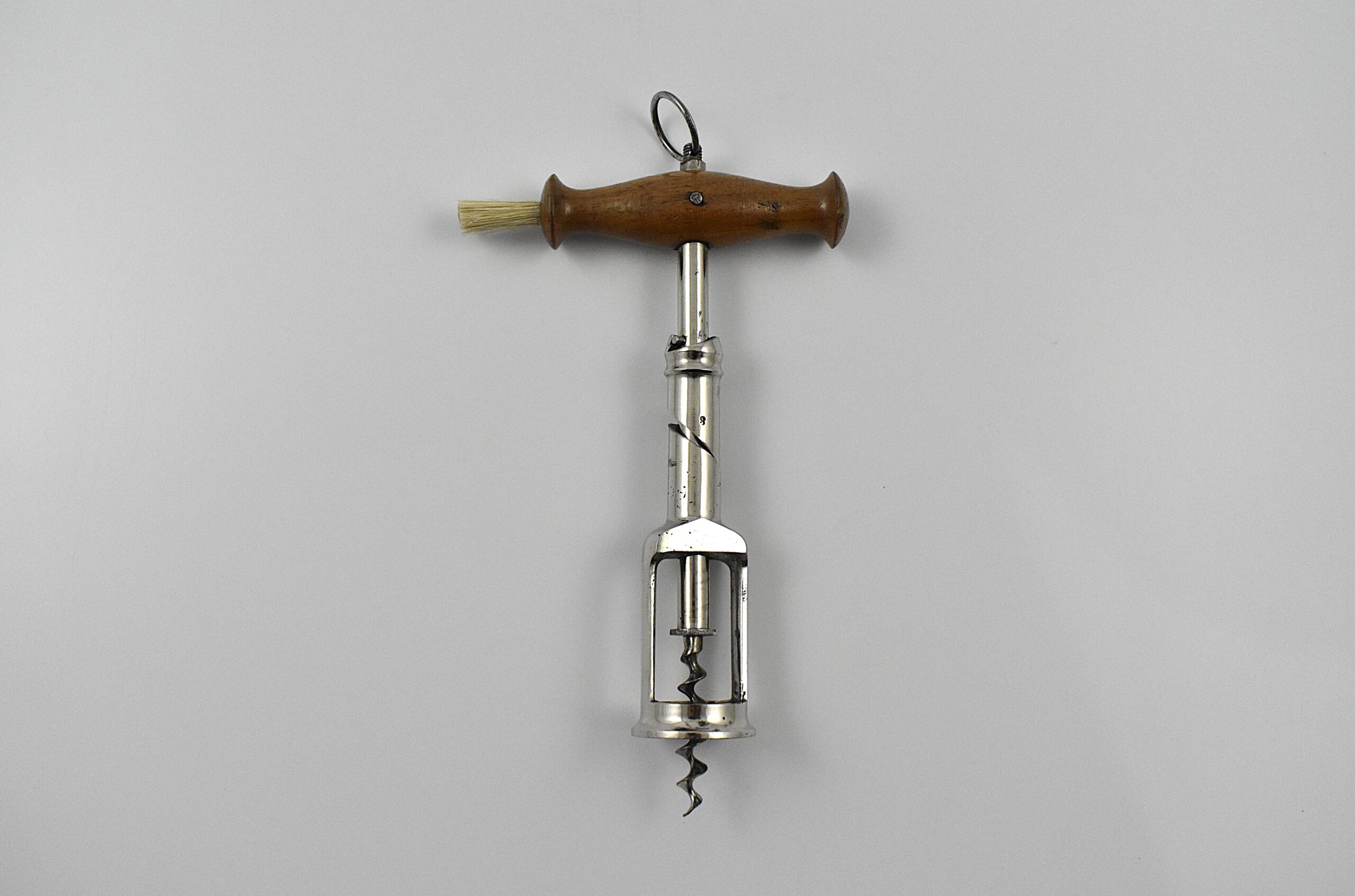 A German chrome plated steel open frame corkscrew with turned wood handle incorporating a dusting brush and hanging ring. The handle is turned into the cork and when the two pins in the shaft engage the top of the second shaft, this reverses and extracts the cork from the bottle. This design originates from the 1880 German patent taken out by Freidrich Kummer and later adapted by his son Louis Kummer. Perfect centre cut bladed worm.